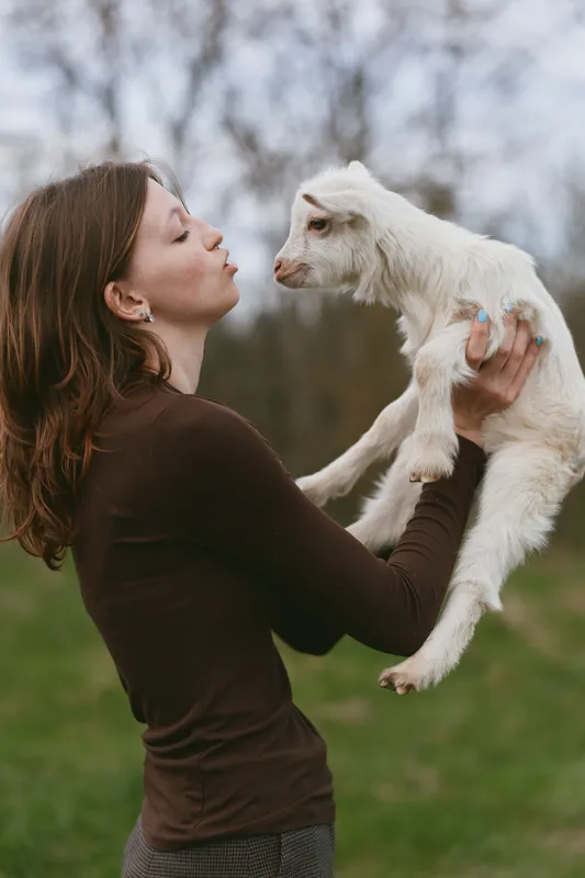 A woman in a brown top lifts a white goat kid in a sunlit field, sharing a tender moment. A candid portrait of outdoor lifestyle, animal companionship, and gentle affection.
