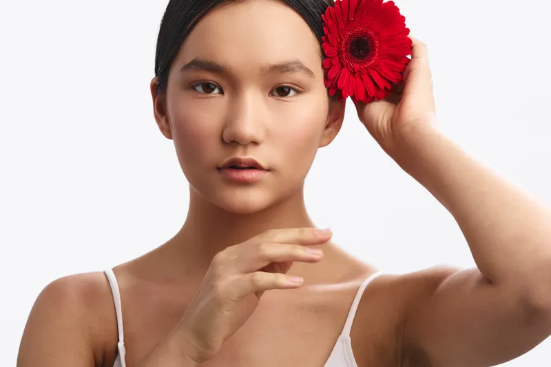 Asian woman holds a red flower near her head in a close studio portrait. Soft lighting highlights her calm, confident expression, creating an elegant, intimate mood.