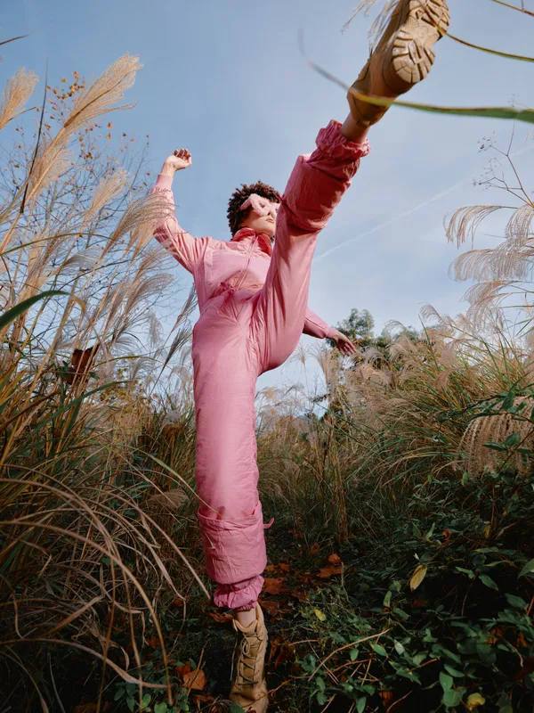 Outdoor scene of a woman in a pink jumpsuit jumping through tall grasses, capturing dynamic movement, energy, and a sense of freedom in bright natural light with vivid colors and joyful mood