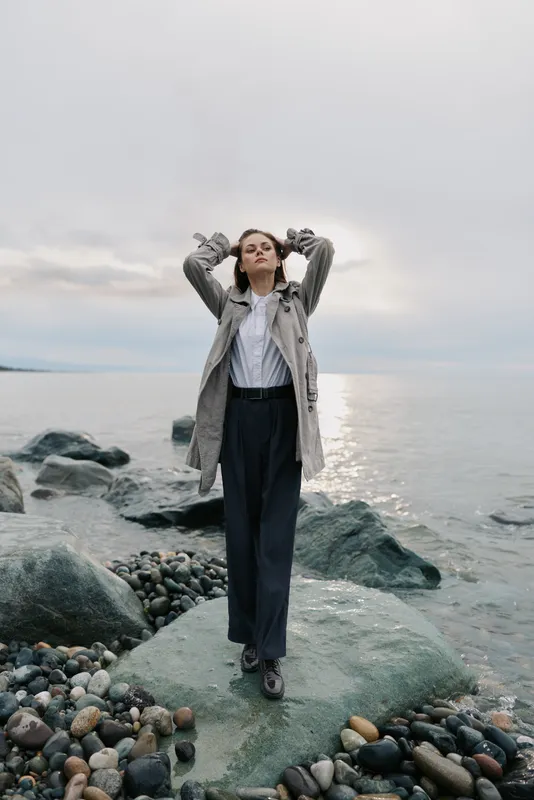 Young woman in coat and trousers stands on rocky shore with arms raised, eyes closed, facing calm sea under cloudy sky. Concept of freedom, nature, and peaceful moment outdoors.