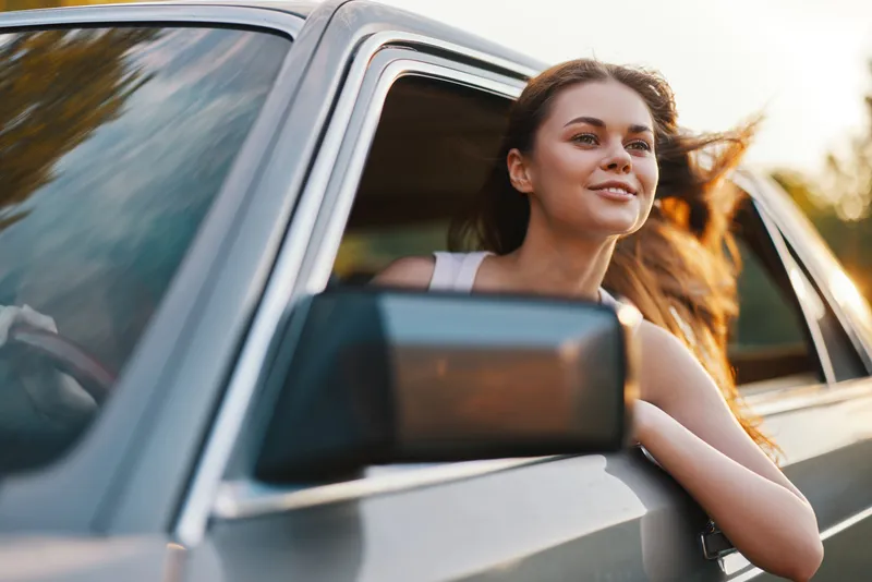 Young woman enjoying the breeze in a car, with long hair flowing outside the window, exuding joy and freedom during a sunny day.