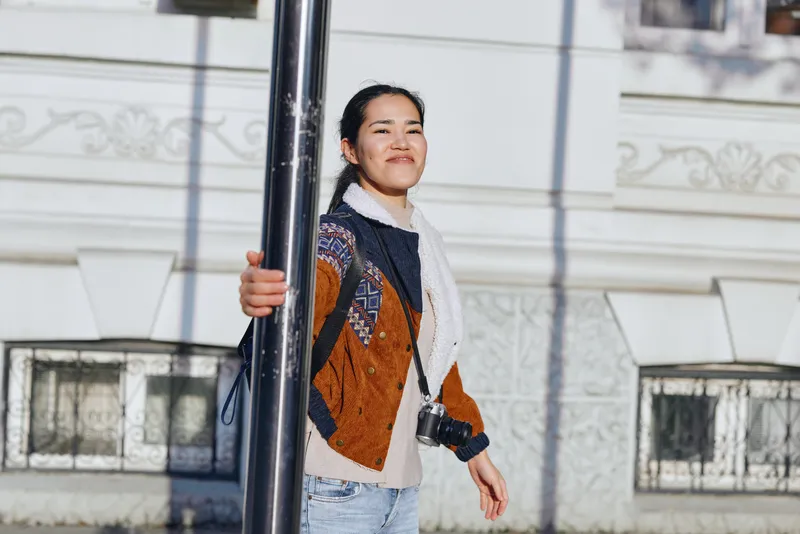 Woman smiling near a street pole, camera in hand, urban outdoor fashion scene with patterned sweater and scarf, bright afternoon light and casual vibe.