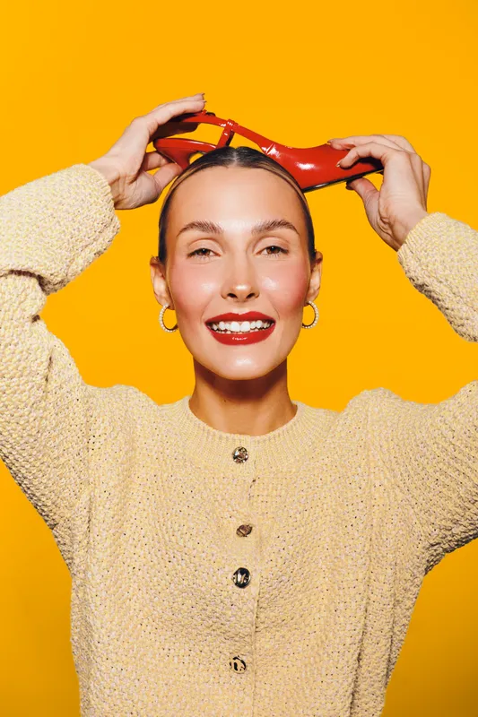 Smiling woman in beige sweater holds red high heel shoes above head on yellow background. Concept of fashion, style, happiness, and bright colorful portrait.