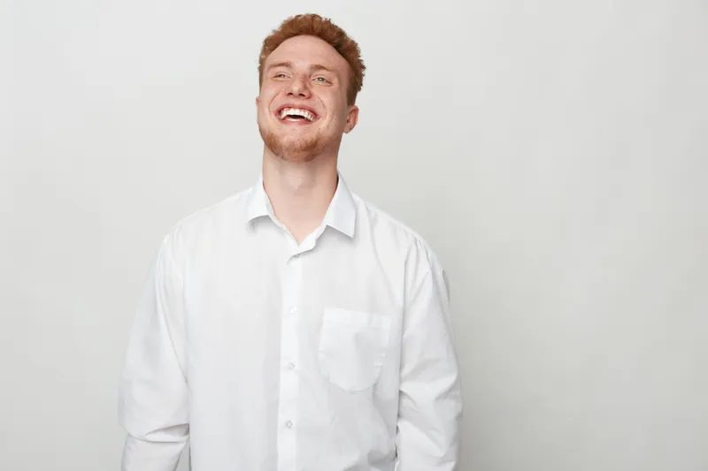 Happy young man with red hair and beard wearing white shirt laughs joyfully against plain light background. Concept of positive emotions, happiness, and casual lifestyle.