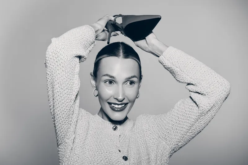 Smiling white woman with light skin and brown hair holds black high heel shoe above head, wearing knitted sweater. Concept for fashion, style, and joyful mood in studio portrait.