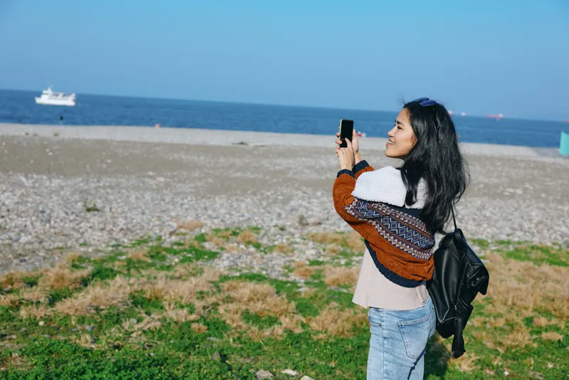 Woman on a sandy beach holds a smartphone, capturing a seascape with blue sky, casual cardigan, jeans and backpack, highlighting outdoor photography, travel, coastal leisure and natural light