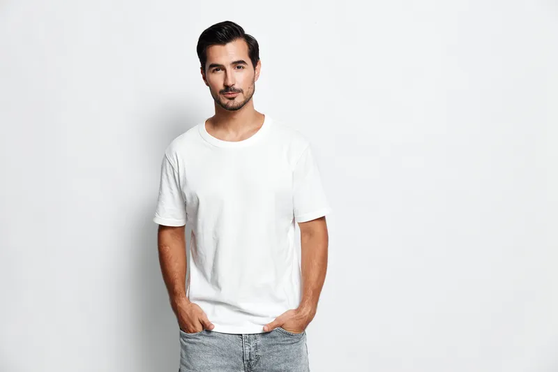 Confident young man standing with hands in pockets, looking at the camera, wearing a casual white t-shirt and jeans, against a plain solid background. Style concept.