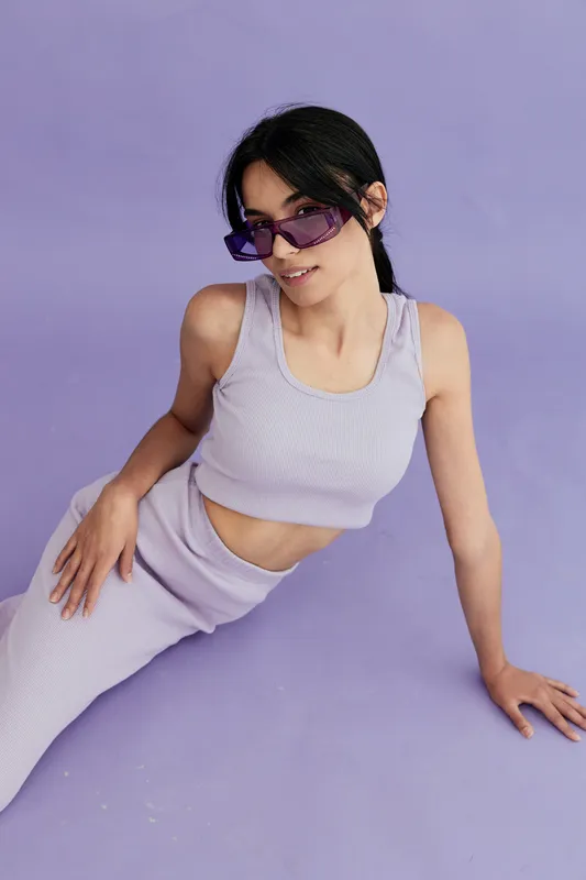 Young woman wearing purple sunglasses and casual ribbed outfit relaxing on lavender background, stylish and modern look with soft monochromatic tones.
