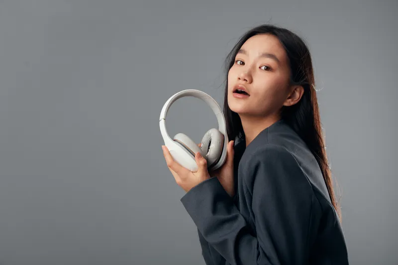 Woman holding headphones with hand in studio, looking at camera with slight smile, dressed in dark jacket on gray background, technology and music accessories concept with relaxed expression.