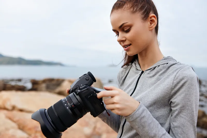 Young woman photographing on a rocky beach, wearing a gray hoodie and smiling, enjoying nature and photography amidst calm ocean waves