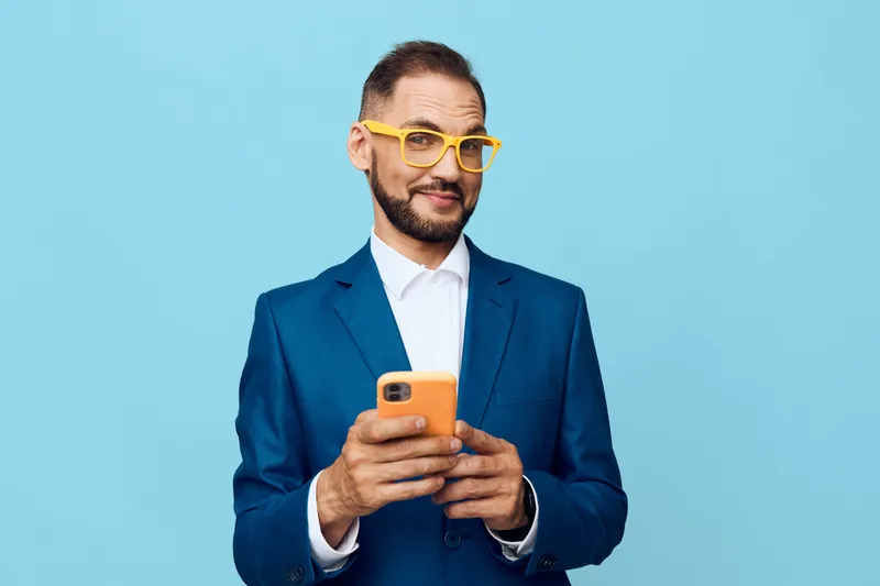 A professional businessman stands against a solid colored backdrop, projecting confident emotion while checking messages on a smartphone, portraying focus, poise, and approachable energy in a modern
