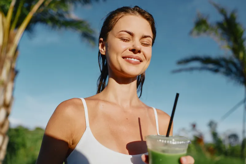 Cheerful woman outdoors, smiling with eyes closed, sunlit moment, refreshing drink in hand, casual tank top, warm breeze, nature, greenery, tropical backdrop