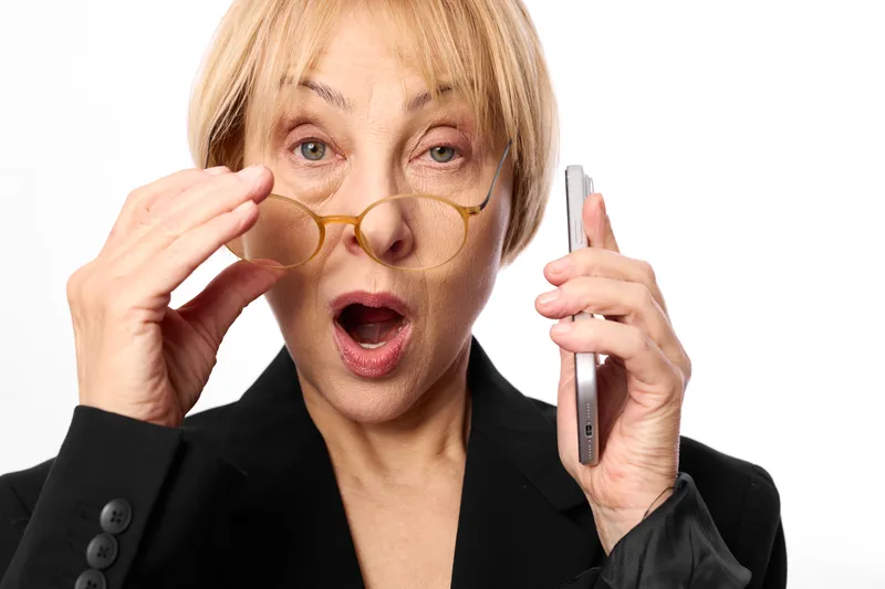 Surprised woman holding glasses and phone in studio white background, blonde hair, business suit, expressive face, communication moment, looking at camera today