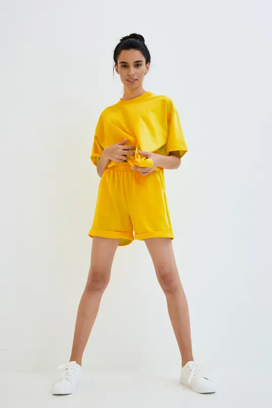 Young woman in bright yellow casual outfit posing confidently against white background, showcasing modern summer fashion and relaxed style with white sneakers.