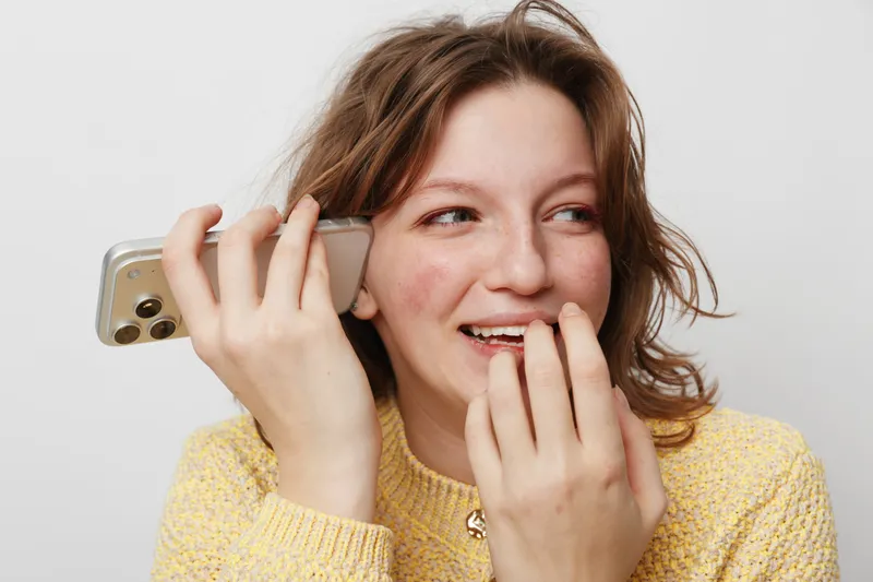 Young woman with natural skin and brown hair smiling while holding smartphone near ear. Casual lifestyle concept with communication, happiness, and candid moment on white background.