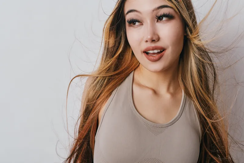 Young woman with long hair wearing sleeveless top looking aside with slight smile on light background, casual portrait with natural makeup and relaxed expression.