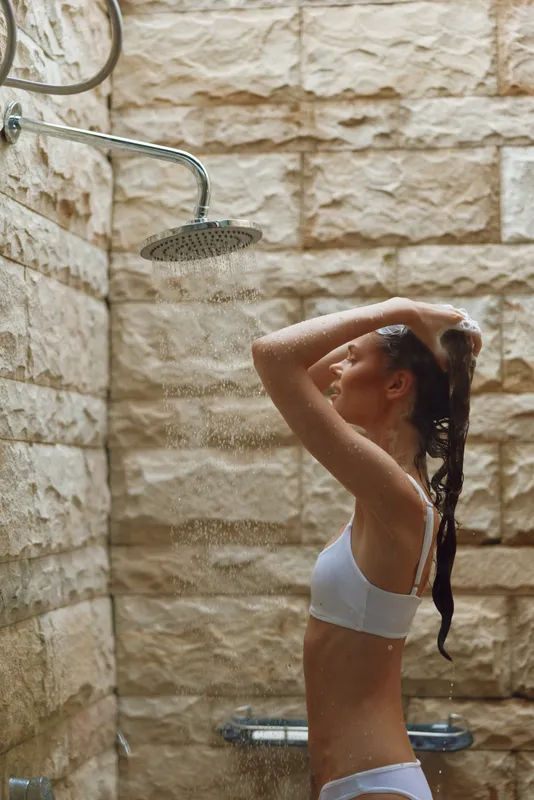 woman showering outdoors in white underwear under rain showerhead with stone wall background, natural light and refreshing water spray on skin