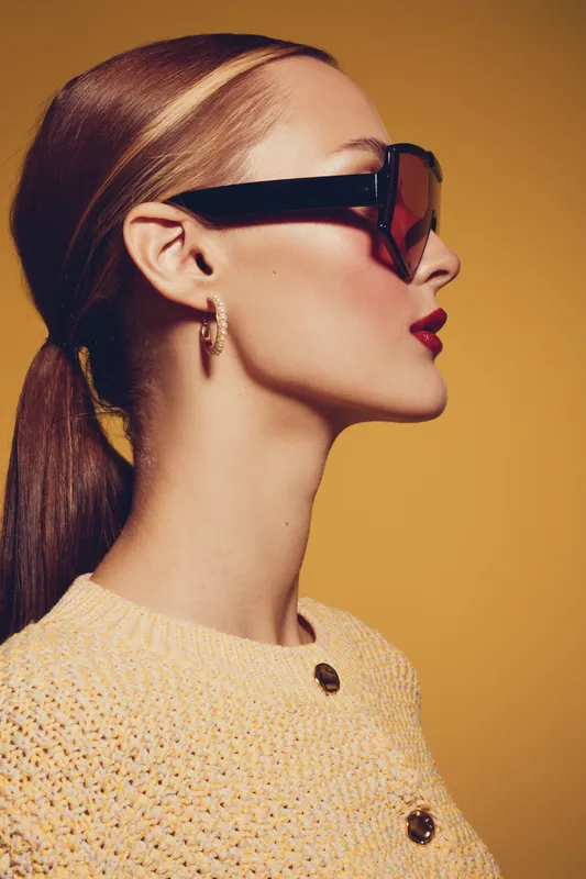Profile of stylish woman with sleek ponytail wearing large sunglasses and red lipstick against yellow background. Concept of fashion, beauty, elegance, and modern accessories.