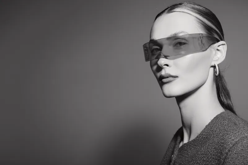Caucasian woman with fair skin wears futuristic sunglasses and a textured jacket against a grey background. Concept of fashion, style, and modern accessories.