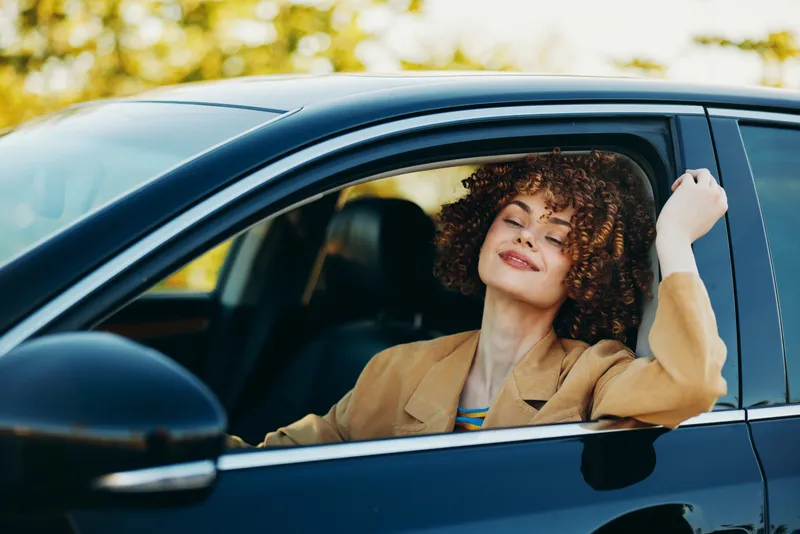 A cheerful woman drives a car with a relaxed smile, natural light in the cabin, and a retro film vibe. Concept for lifestyle joy and travel in a candid automotive moment.