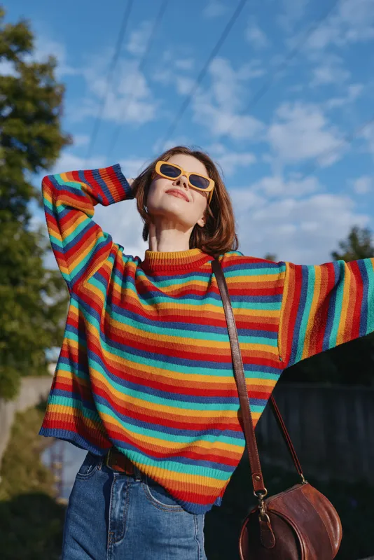 woman in her 30s wearing a rainbow sweater and yellow sunglasses, smiling with arms raised outdoors on a sunny suburban street, casual jeans and brown crossbody bag, joyful travel vibe