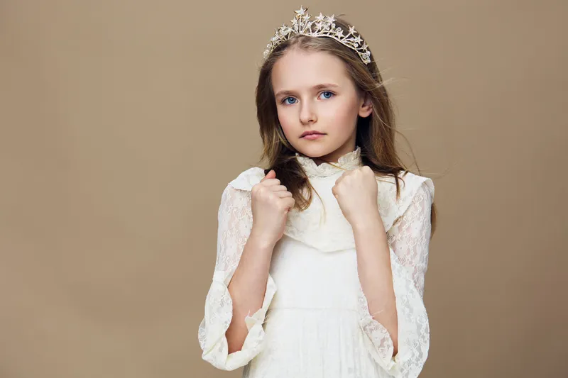 Young girl in a white dress with a crown, looking serious and elegant against a soft beige background, reflecting innocence and royal charm