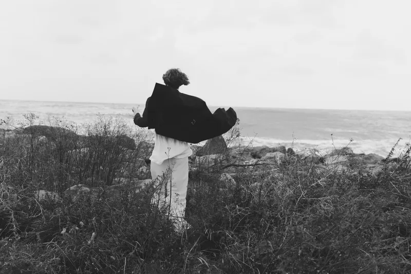Embrace on a windswept coastline, a cloaked figure lifting another along a monochrome shore, conveying care, resilience, and loss with a dramatic silhouette against the sea, a cinematic moment for