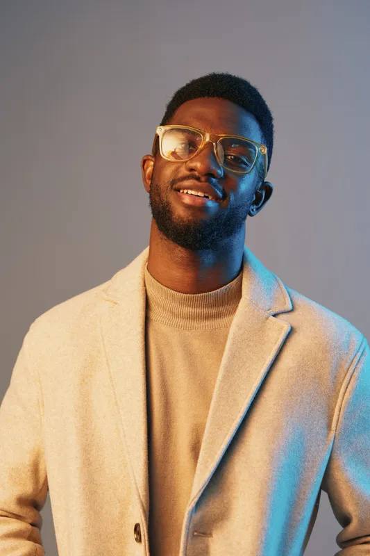 Stylish black man in eyeglasses smiling confidently against a blue and beige background, wearing a sweater and a coat, showcasing urban fashion and self assurance