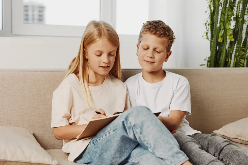Two children sitting on a beige sofa indoors, girl writing on a clipboard and boy watching attentively. Concept of friendship, childhood learning, and collaboration.