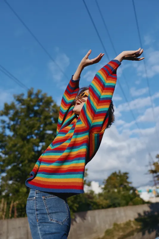 person in a rainbow sweater stretching outdoors with sunglasses under a blue sky, wearing jeans and joyful expression, colorful knitwear, sunlight and casual lifestyle feeling of freedom.