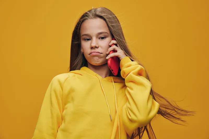 Girl in yellow hoodie on phone with serious expression standing against yellow background, long hair flowing and casual style for communication and technology.