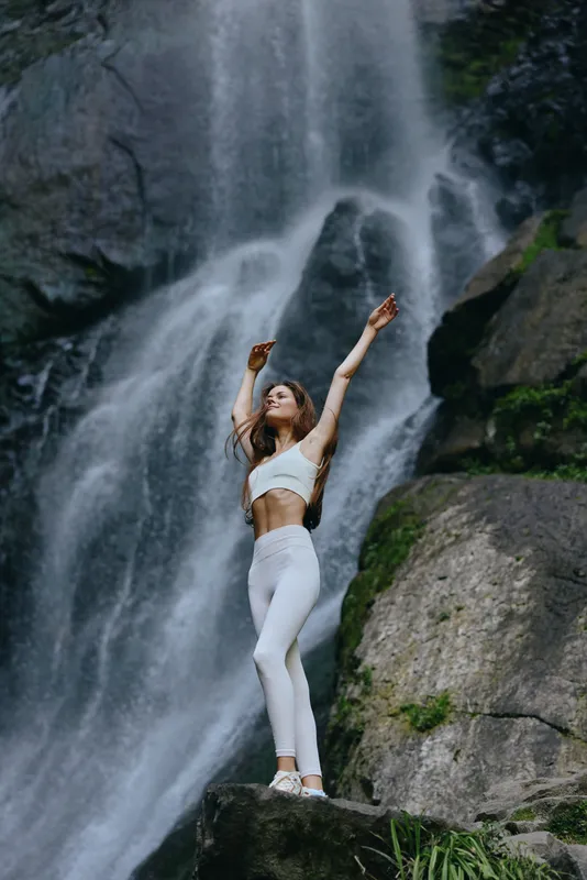 Young woman practicing yoga in white activewear on rocky terrain near a powerful waterfall, showcasing balance, nature, and serene outdoor meditation.