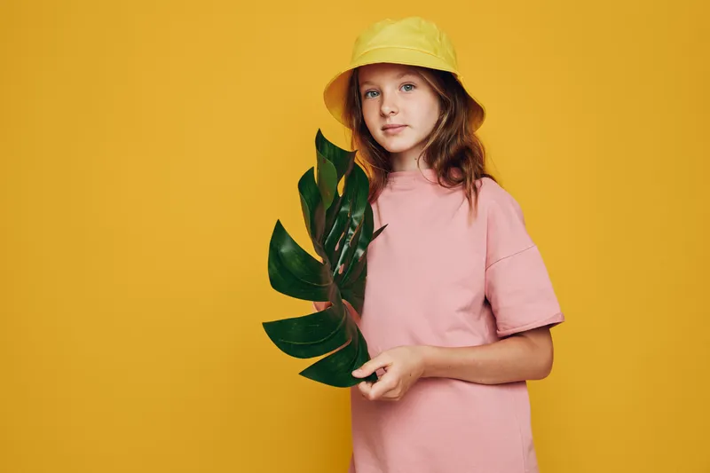 Young girl in a yellow hat holds a large green leaf wearing a pink t-shirt on a yellow background.