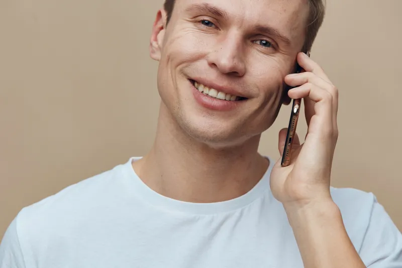 Smiling man talking on mobile phone with friendly expression, casual white t-shirt, blonde hair, light skin, indoor background, close-up shot, cheerful and relaxed mood, lifestyle concept