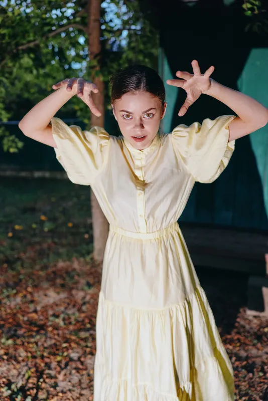 Playful woman in a pale yellow dress poses outdoors in a park, raising hands with a surprised expression, capturing a candid moment of whimsy and energy.