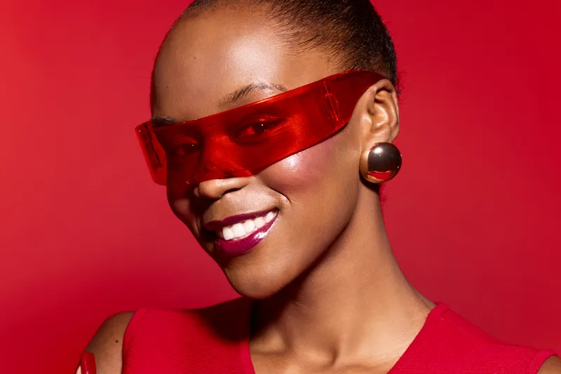 Smiling African American woman with dark skin wearing red futuristic sunglasses and red outfit against red background. Concept of fashion, style, confidence, and modern accessories.