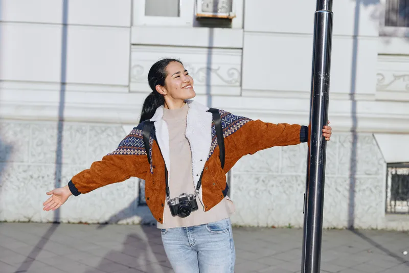 Woman outdoors with open arms and joyful smile beside a street lamp, casual jacket and jeans, expressing freedom, happiness, social connection, and urban lifestyle in an inviting city environment