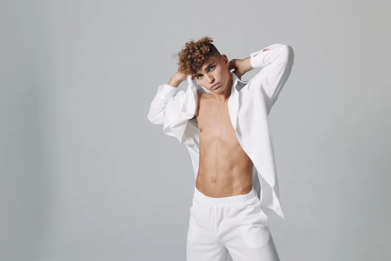 Young man with curly hair posing in white open shirt and pants against gray background, showing toned abs and confident expression in studio portrait.