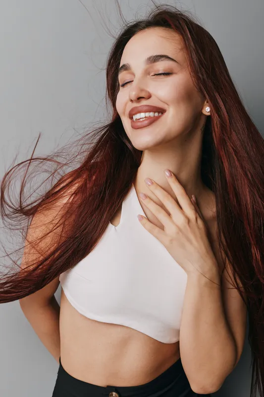 Young woman with long hair smiling with eyes closed wearing white crop top and black pants against gray background. Happiness, beauty, casual style and natural makeup closeup portrait.
