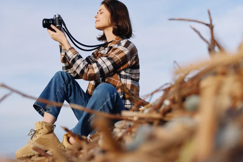 Photographer woman in plaid shirt and jeans sits on a hillside, using a camera to capture landscape and scenery amid tall grass, blue sky, warm daylight and tranquil horizon