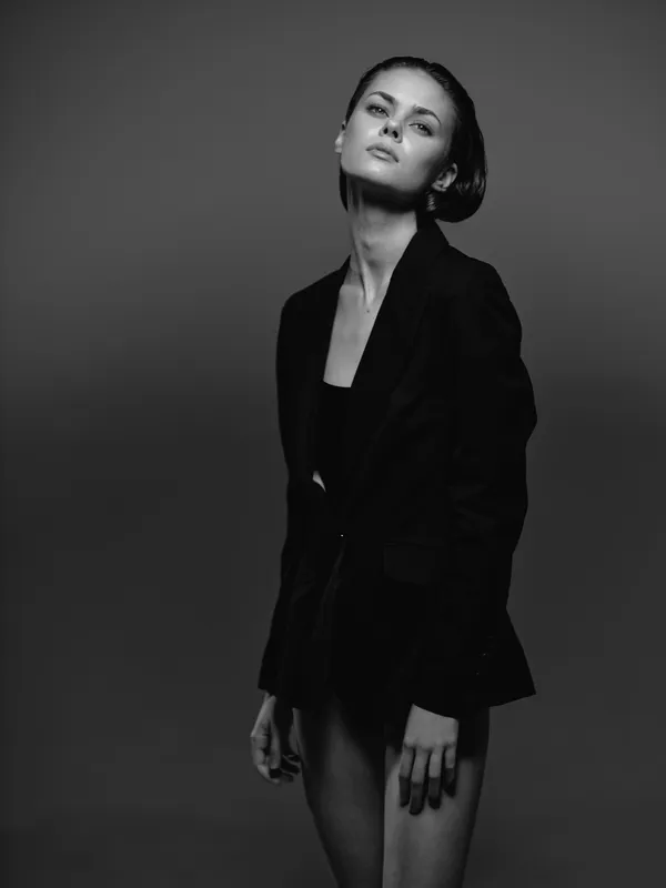 Black and white portrait of a confident woman in a blazer with stylish makeup and wet hair, posing elegantly in a dark studio setting.