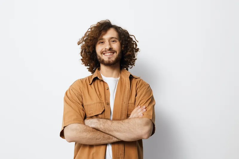 Smiling young man with curly hair wearing a brown shirt, standing confidently with arms crossed against a light background