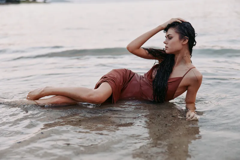 Model lies semi-submerged on a sandy shore, wearing a burgundy dress. Wet hair clings to her skin as she rests, eyes closed, radiating calm and allure.