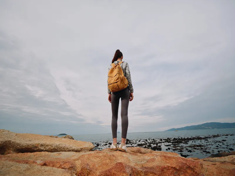 Young woman with backpack enjoying a coastal view, wearing casual outdoor clothing, representing adventure and exploration against a serene ocean backdrop