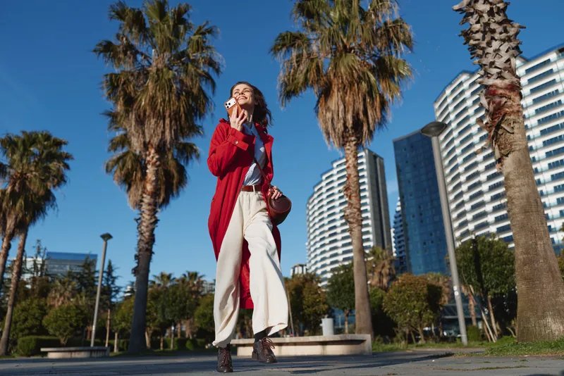 Young woman in a red long cardigan walks through a sunny urban park, enjoying a bite of ice cream as tall palm trees frame modern buildings and a clear blue sky.
