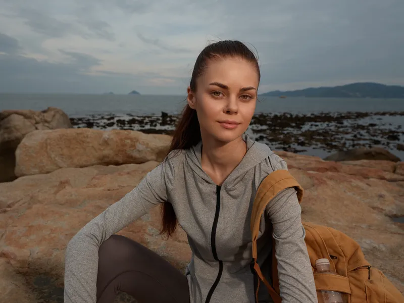 Young woman in athletic wear enjoying a moment by the seaside, showcasing a serene expression against a rocky background with soft, muted colors