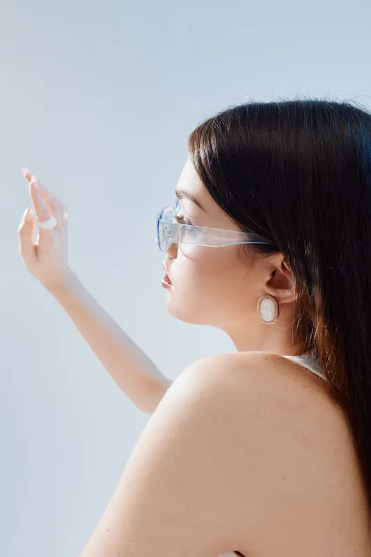 Young woman with long dark hair wearing transparent glasses and earrings looks thoughtfully into the distance. Soft natural light highlights her beauty and fashion style.