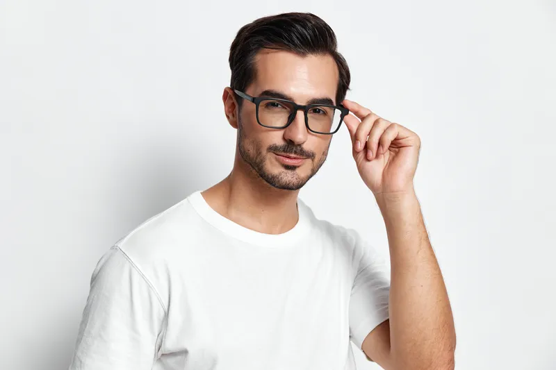 Stylish young man with glasses poses confidently, wearing a white t-shirt and smiling at the camera, isolated on solid light background. Fashion concept.