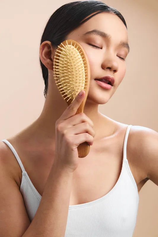 Asian woman gently brushes her hair with a wooden paddle brush, eyes closed and calm, capturing a serene personal care moment in a soft minimalist setting.