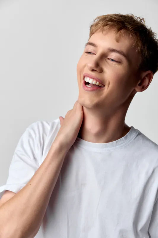 Smile, neck touch, casual, young man, white shirt, happiness, relaxed, candid, studio shot with soft lighting and clean background conveying warmth, confidence and approachable charm